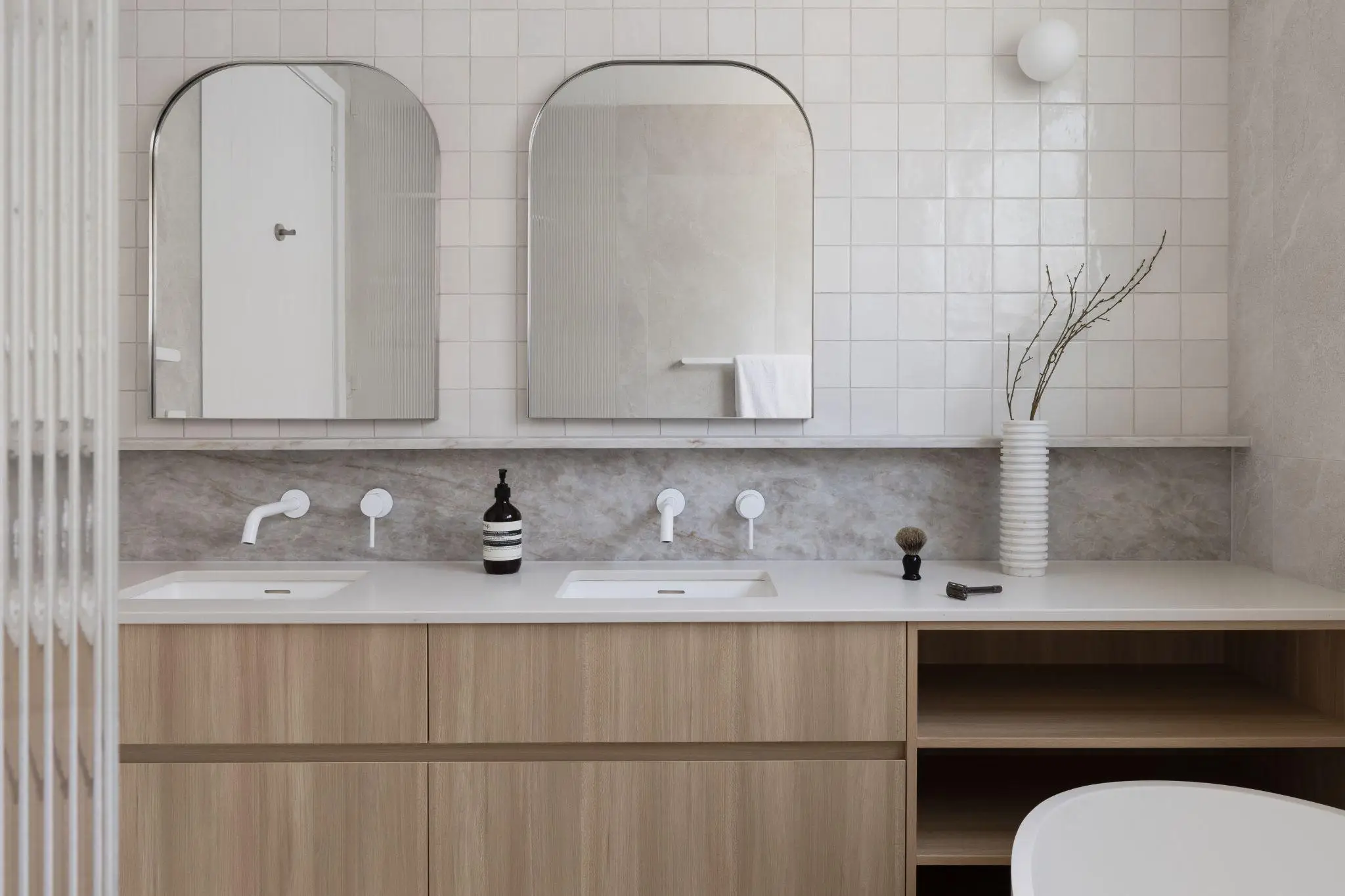 Modern bathroom with dual sinks, wooden cabinetry, arched mirrors, and ceramic vase on tiled wall
