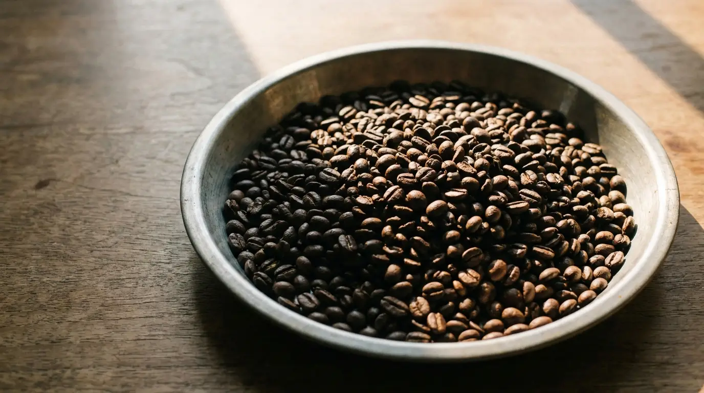 Metal bowl filled with roasted coffee beans on wooden surface in natural light