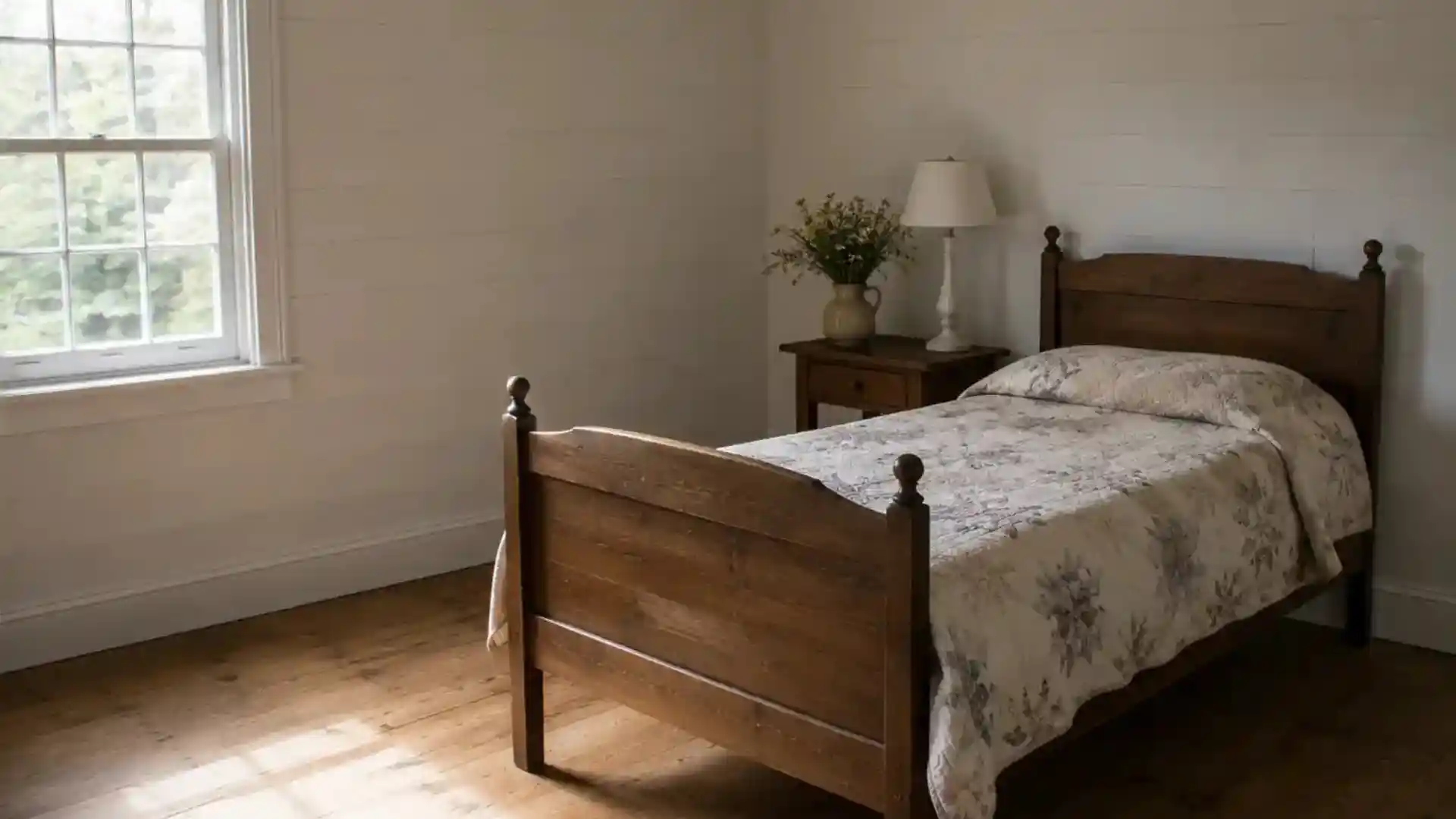 sparsely furnished cottage bedroom with one quilt, lamp and vase of flowers
