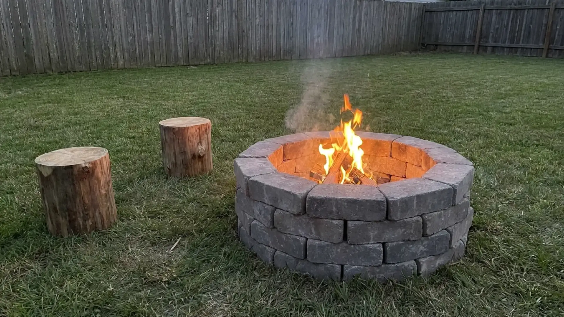 stone ring firepit with burning fire and log stump seats on green lawn