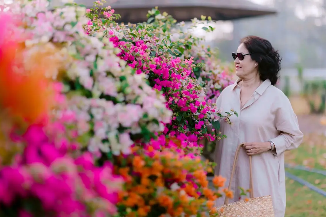 Woman in sunglasses admires vibrant bougainvillea flowers outdoors, holding a woven bag