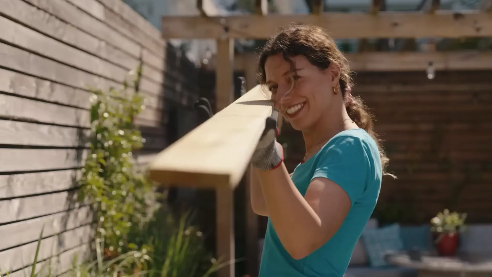 woman carrying wooden plank wearing work glove while smiling during outdoor diy project