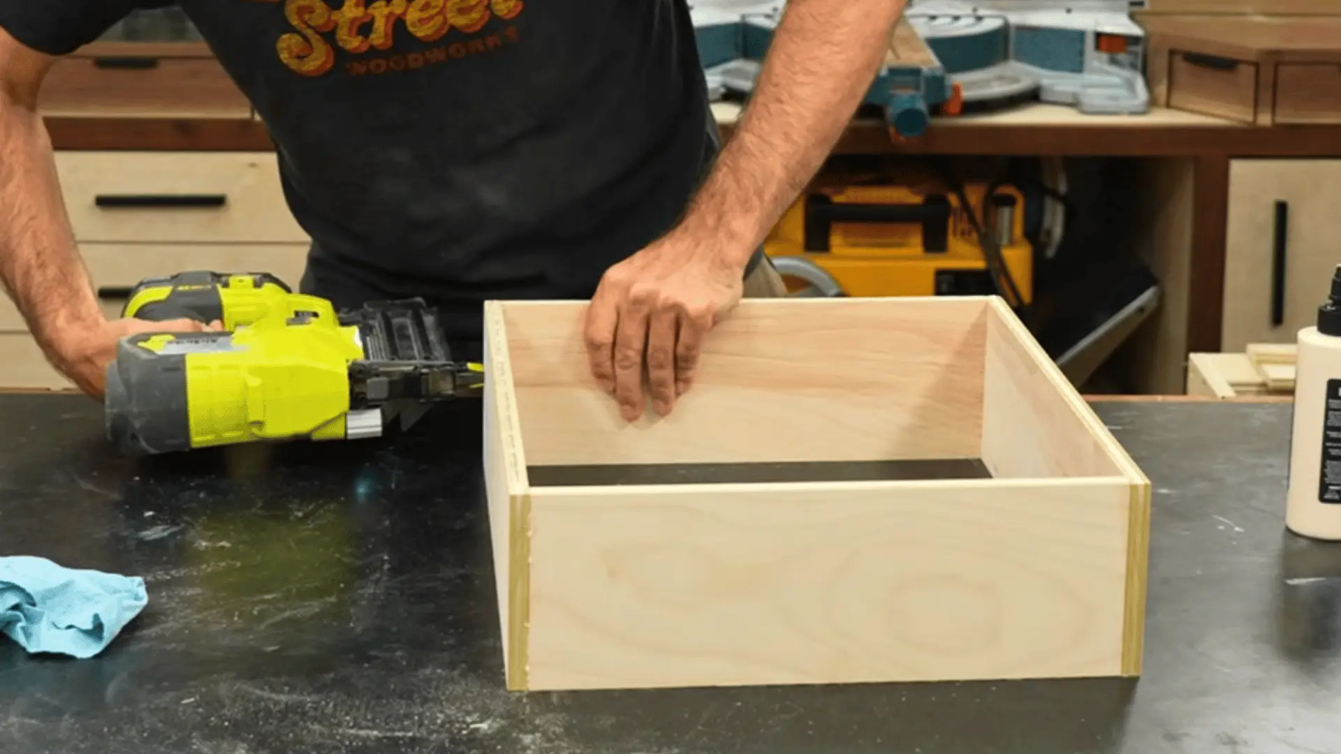 woodworker assembling a plywood drawer box on a workbench with a brad nailer nearby