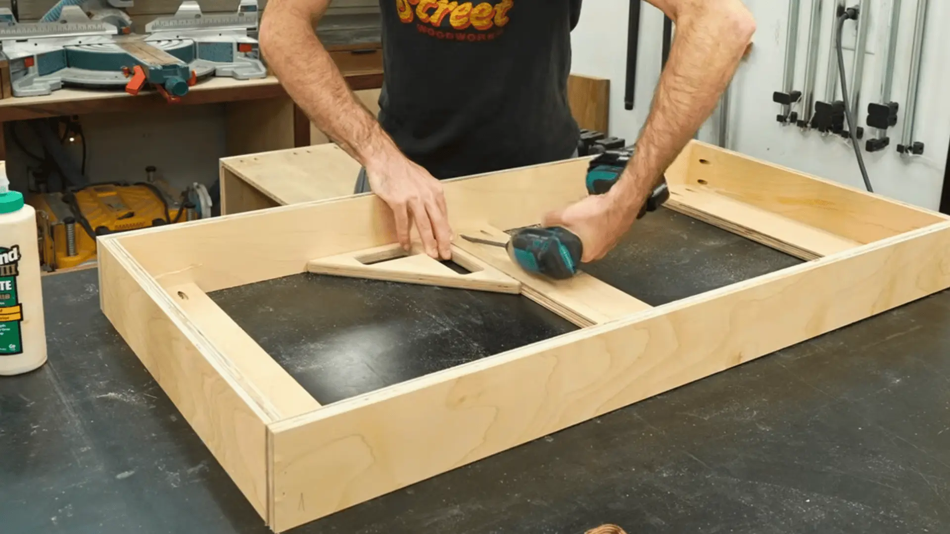 woodworker assembling a plywood toe-kick frame with a drill, using a triangular brace to hold the corner square