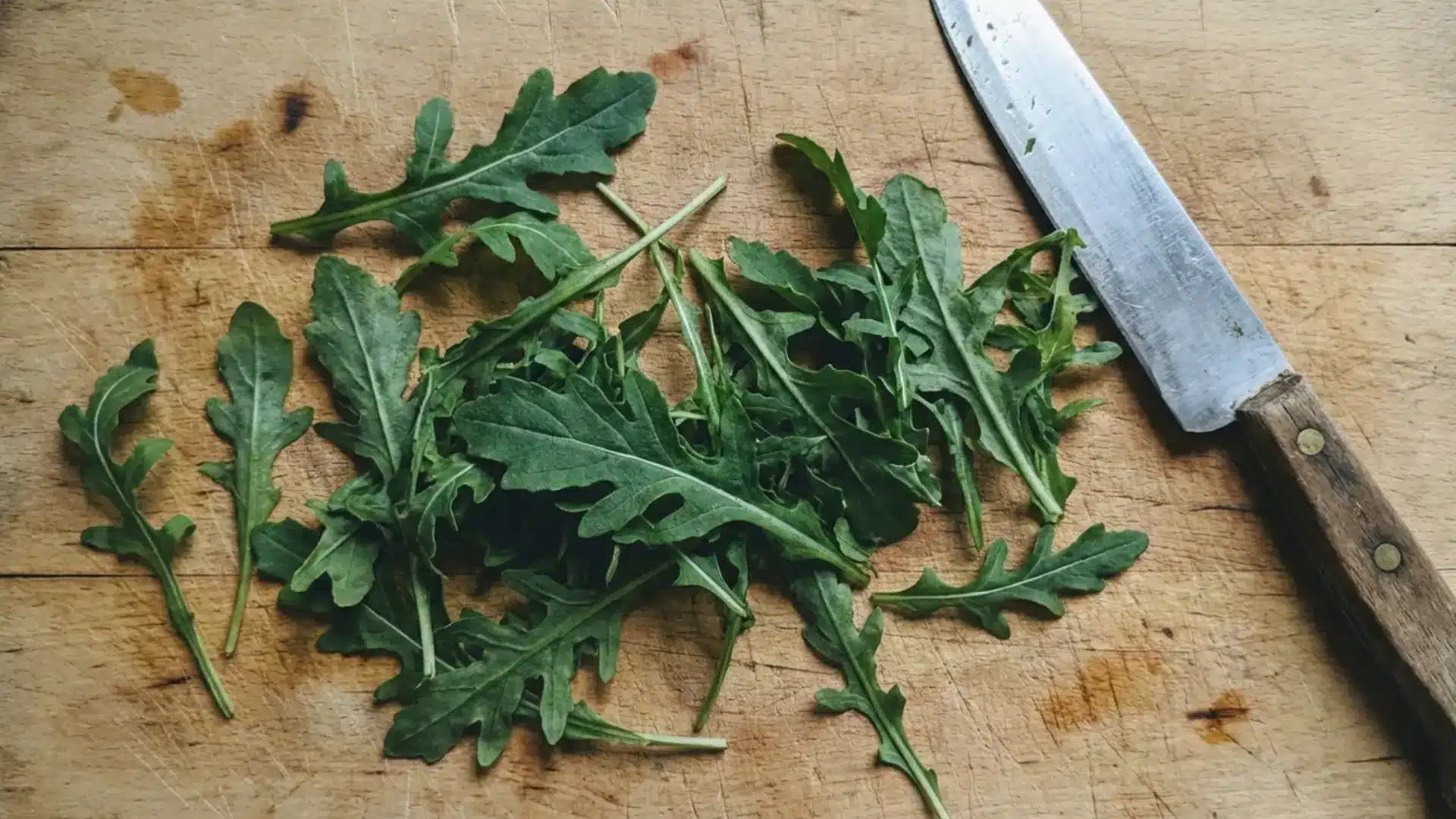 fresh arugula leaves spread on a wooden cutting board beside a kitchen knife