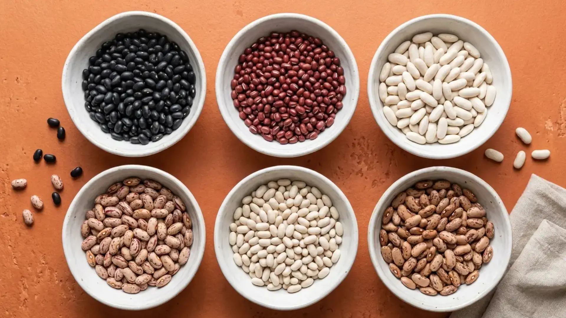 six bowls of different dry bean varieties kept in small white bowls on an orange background