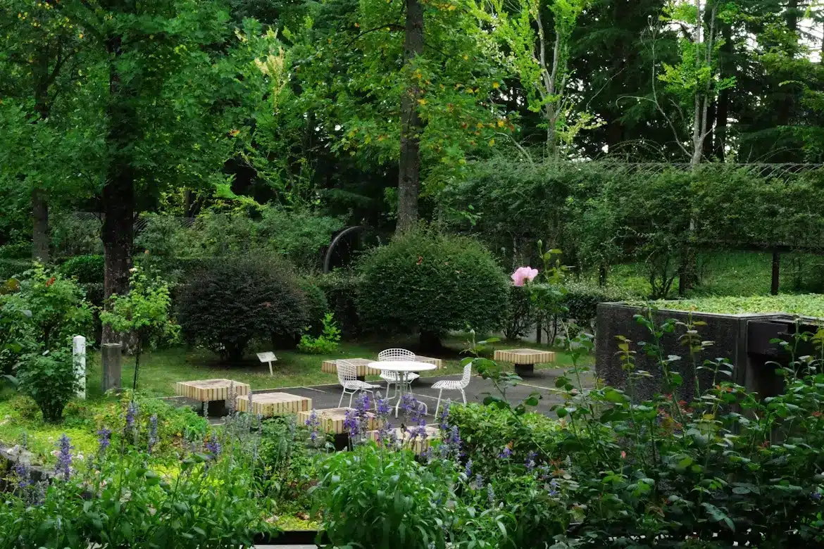 Outdoor seating area with white metal chairs and wooden benches surrounded by lush greenery