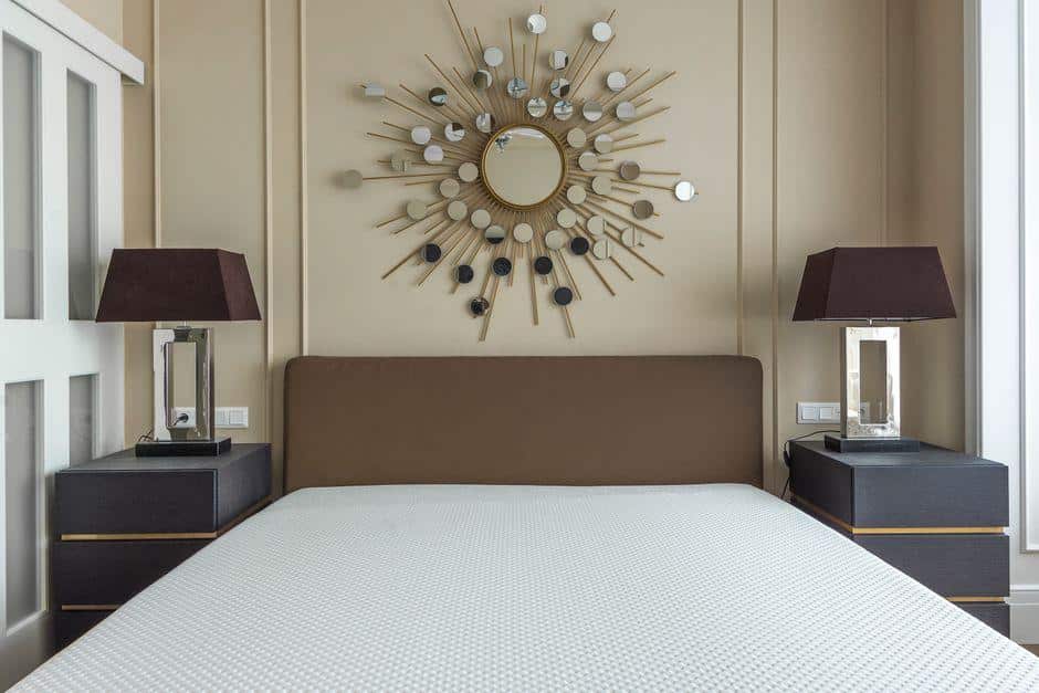 Modern bedroom with sunburst wall decor, two dark bedside tables with lamps, beige tones