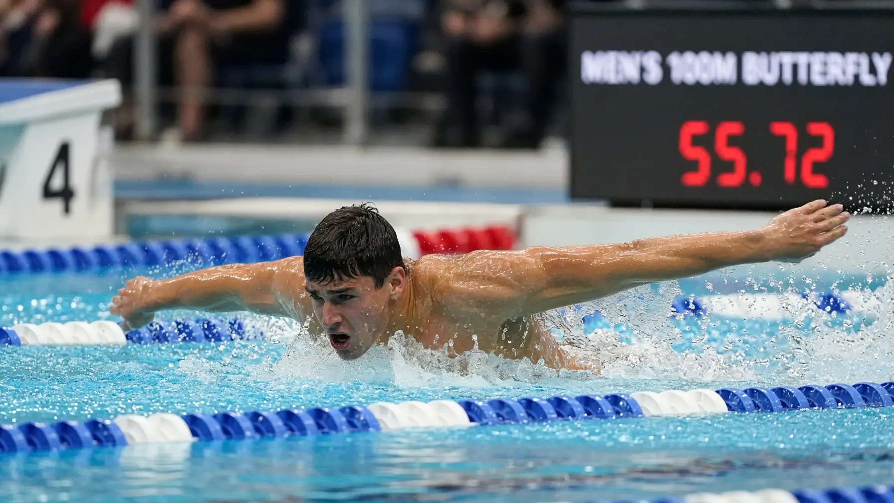 Swimmer performing butterfly stroke in indoor pool with timing board in background