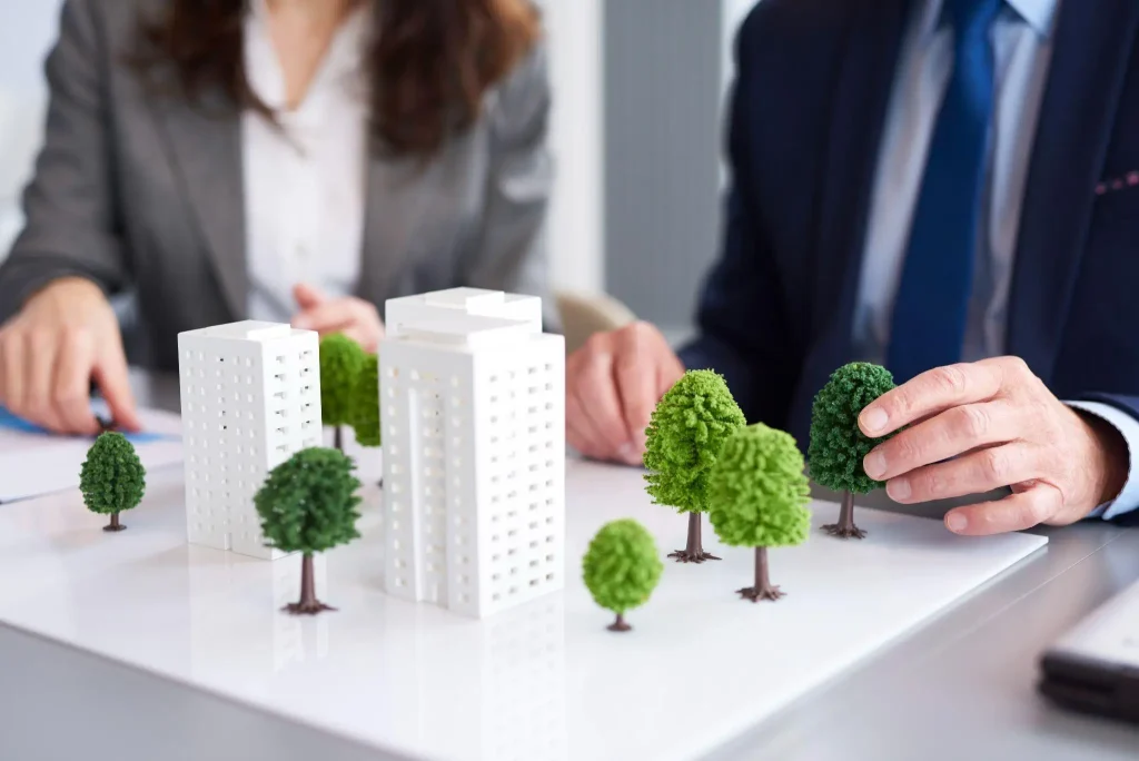 Miniature architectural models of buildings and trees on a desk with two people discussing plans